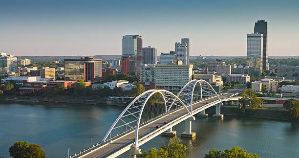 Drone shot of downtown Little Rock, Arkansas during golden hour, featuring the distinctive white arch bridge spanning the Arkansas River. The city skyline includes modern high-rise buildings and office towers rising above the riverfront district. Lush green trees line both sides of the river, with residential and commercial areas extending into the distance. The warm afternoon light illuminates the urban landscape and creates reflections on the calm river water. • Authorization was obtained from the FAA for this operation in restricted airspace.