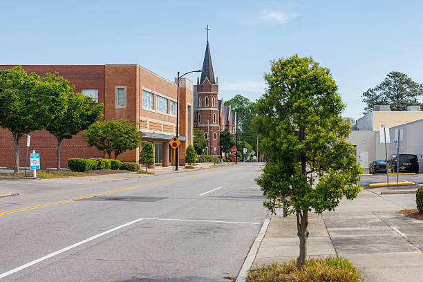 A quiet two-lane road runs through downtown Wilson, North Carolina on a clear spring morning. Red brick commercial and institutional buildings line the left side of the street, flanked by trimmed green trees and low hedges. A parking sign for a law office stands near the curb on the left. At mid-block, a Gothic Revival brick church with tall pointed steeple topped by a cross rises prominently above the surrounding buildings, its arched windows and ornate tower visible against a pale blue sky. A small young tree grows from the sidewalk in the right foreground, with parked vehicles and a low commercial building visible on the right side. Stop signs and yellow warning signs mark a cross street intersection further down the road.