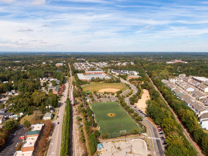 High-resolution drone images capture the small-town charm of Apex, North Carolina, showcasing its tree-lined neighborhoods, downtown streets, and surrounding green landscape on a bright, clear day. The photos highlight the mix of residential areas, local businesses, and community landmarks, including the town’s water tower and historic district, under a vivid blue sky.