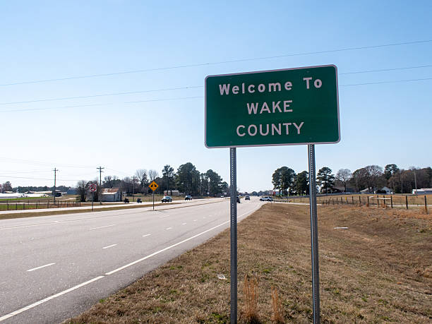 This horizontal shot captures a standard green rectangular highway sign with white lettering that reads "Welcome To WAKE COUNTY." The sign is mounted on two metal posts on the shoulder of a road in North Carolina. The perspective is from a low angle, emphasizing the sign against a vast, clear blue sky. In the background, a rural scene unfolds with dry winter grass, a white wooden farm fence, a few scattered evergreen and deciduous trees, and a distant house. The lighting is bright and direct, highlighting the reflective texture of the municipal signage.
