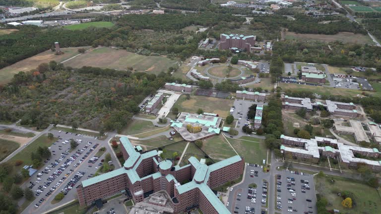 Aerial drone view of a large medical campus in Brentwood, Suffolk County, Long Island, NY, showcasing multi-wing architecture, structured parking, open grounds, and surrounding landscape in clear day