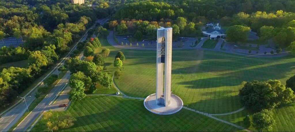 Aerial view of a large green park with a tall concrete memorial bell tower at the center, surrounded by walking paths, trees, and nearby buildings and roads.