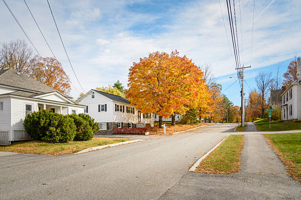 White wooden detached houses along a tree lined street in a mountain village on a sunny autumn morning. A beautiful orange maple tree stand in front of one of the houses. Bethel, ME, USA