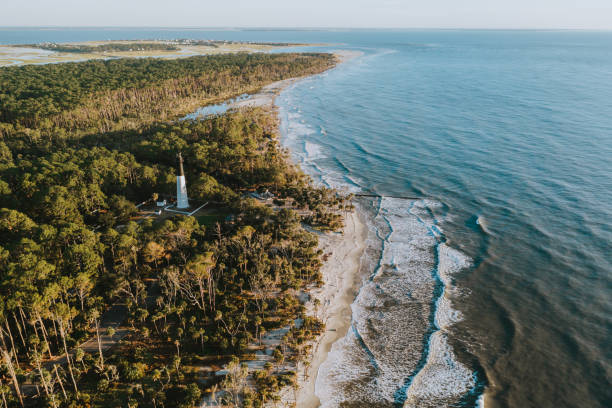 Aerial view of a South Carolina barrier island with a lighthouse, maritime forest, and waves rolling onto the beach.